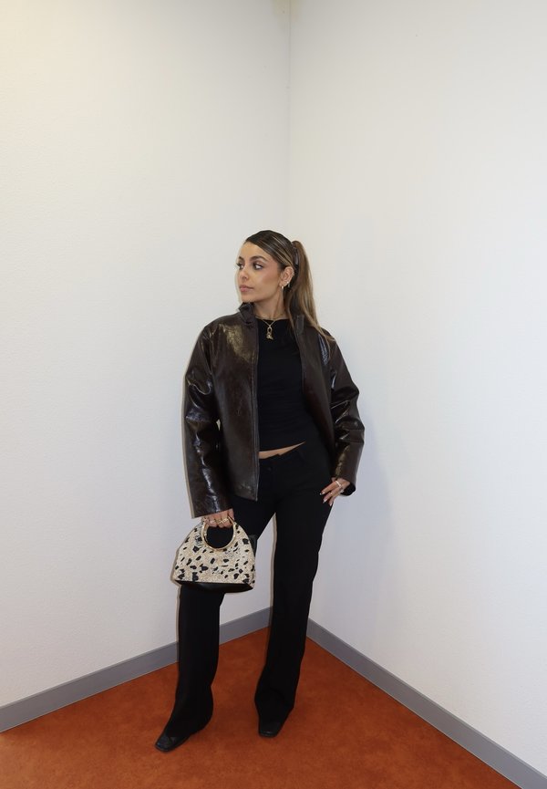 Jeune femme en tenue noire et veste en cuir marron tenant un sac à main à motifs, debout dans un coin avec des murs blancs et un sol orange.