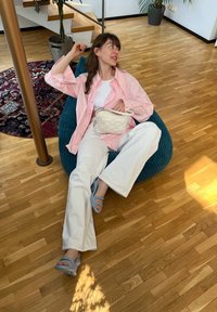 Calças largas brancas, camisa rosa de botões, relaxando sobre uma pufe de ribana teal. Apresentando uma bolsa branca e sandálias azuis claras com tiras, num chão de madeira.