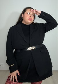 Femme portant un blazer noir avec une ceinture décorative, des collants rouges à motifs floraux et des boucles d'oreilles créoles dorées, tenant une main sur son front tout en regardant sur le côté.