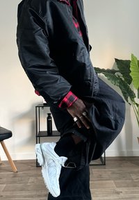 Zwarte bomberjack over een rood-zwart geruite shirt, gecombineerd met donkere broeken en witte sportschoenen. Ringen zichtbaar op de hand.