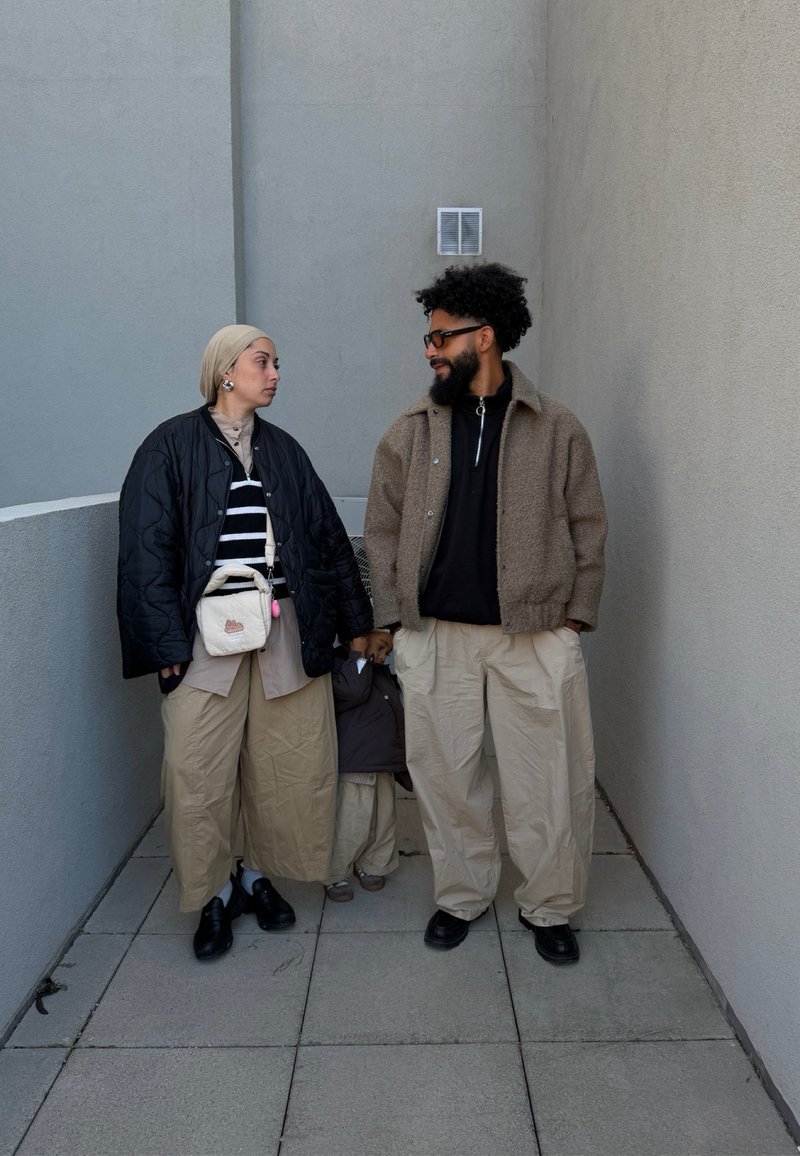 Deux adultes tenant la main d'un petit enfant entre eux, tous vêtus de vêtements superposés aux tons neutres, debout dans un couloir extérieur étroit.
