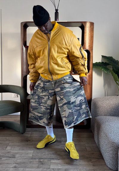 Hombre con chaqueta amarilla, gorro negro, pantalones cortos de camuflaje, calcetines blancos Puma y zapatillas amarillas, de pie en el interior cerca de muebles y plantas.