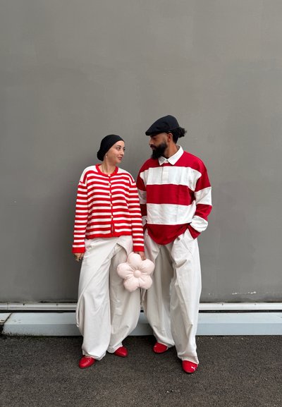 Pareja de pie frente a una pared gris, vistiendo camisetas a rayas rojas y blancas, pantalones anchos blancos, zapatos rojos; la mujer sostiene una almohada en forma de flor rosa.