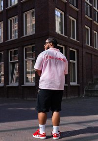 Camisa rosa de manga curta com "CIAO AMORE" em letras vermelhas, combinada com calções pretos e ténis vermelhos, contra um fundo de prédio de tijolos.