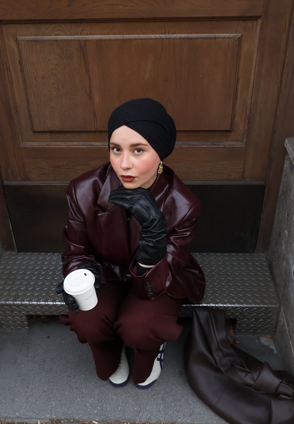 Woman in dark burgundy outfit and black gloves sits on metal step, holding white coffee cup, next to brown leather bag and wooden door.