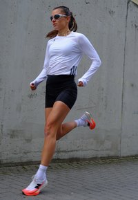 Väljas joostes naine, kellel on seljas valge pika varrukaga särk, mustad lühikesed püksid, valged sokid ja valged jooksujalatsid oranži tallaga.