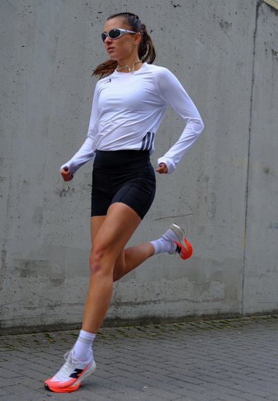 Mujer corriendo al aire libre con camiseta blanca de manga larga, pantalones cortos negros, calcetines blancos y zapatillas de correr blancas con suelas naranjas.