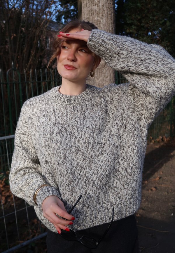 Femme portant un épais pull en tricot gris, se protégeant les yeux du soleil avec une main, tenant des lunettes de soleil noires dans l'autre.