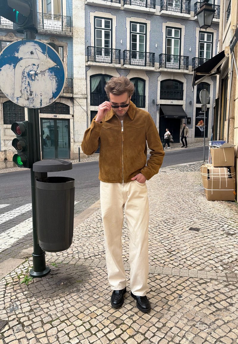 Mann trägt braune Jacke, weiße Hose und schwarze Schuhe, richtet Sonnenbrille auf Kopfsteinpflasterstraße mit historischen Gebäuden und Ampel.