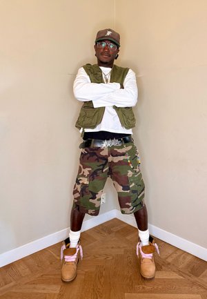 Hombre con gafas, chaleco verde, camisa blanca de manga larga, pantalones cortos de camuflaje, botas color beige con cordones rosas y una gorra oscura, de pie frente a paredes beige.