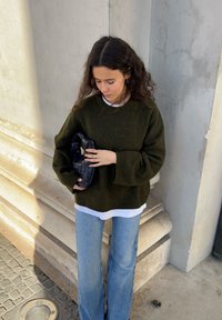 Maglione verde oliva oversize sopra una camicia bianca, abbinato a jeans a zampa d'elefante blu chiaro. Tiene in mano una piccola borsa a tracolla nera intrecciata.