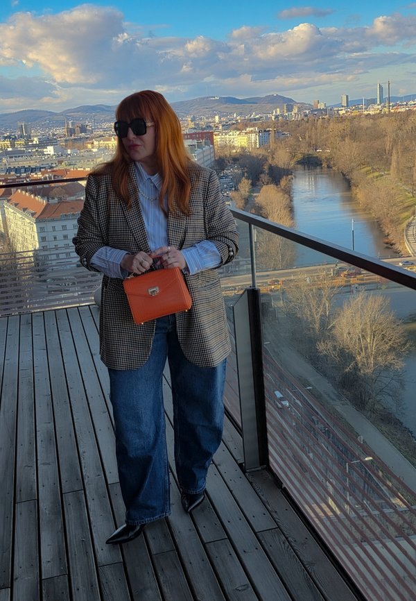 Femme aux cheveux roux portant des lunettes de soleil, un blazer à carreaux, un jean et des chaussures noires à bouts pointus, tenant un sac orange sur un balcon avec vue sur une rivière et le paysage urbain.