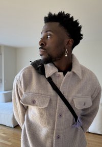 Homme aux cheveux courts et bouclés portant une veste à boutons violette claire et une boucle d'oreille pendante, regardant vers sa gauche à l'intérieur.