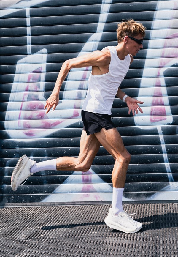 Homme athlétique portant un débardeur blanc, un short noir et des lunettes de soleil courant devant une grille métallique couverte de graffitis en extérieur.