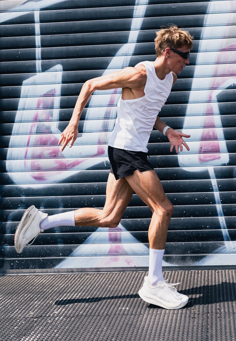Homme athlétique portant un débardeur blanc, un short noir et des lunettes de soleil courant devant une grille métallique couverte de graffitis en extérieur.