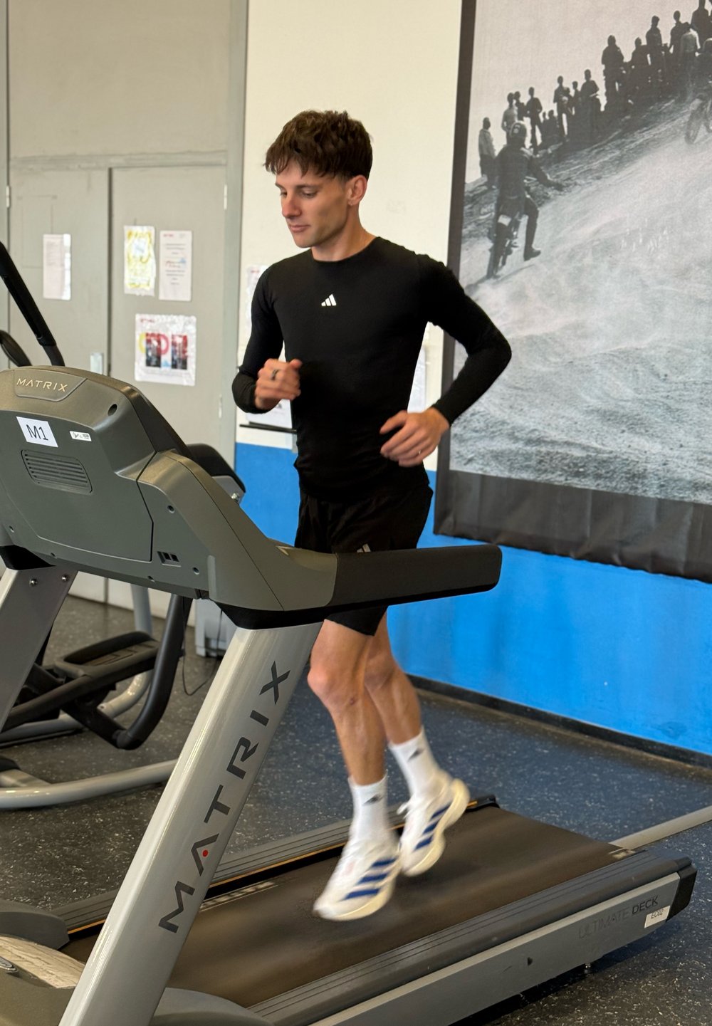 Szürke és fekete kialakítású futópad. Egy fekete hosszú ujjú felsőt és rövidnadrágot viselő személy fut rajta, fehér és kék sportcipőben.