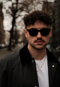 Veste bomber noire avec col en velours, t-shirt blanc en dessous, collier en chaîne dorée et lunettes de soleil noires. Cheveux courts bouclés et moustache.