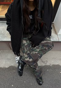 Personne assise à l'extérieur sur une marche, portant un pantalon camouflage, des chaussures Nike noires, une veste noire, une écharpe noire et un t-shirt noir avec un logo NY blanc.