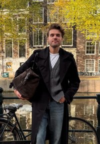 Svart trenchcoat över en grå cardigan och vit skjorta, håller en mörkbrun v�äska. Bakgrunden visar höstlöv och historiska byggnader.