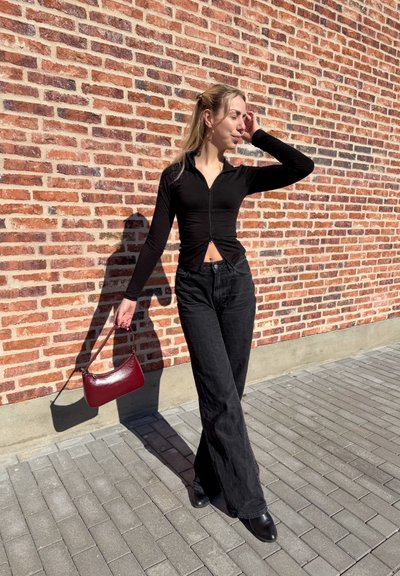 Joven mujer con camiseta negra con cremallera y pantalones anchos negros, de pie contra una pared de ladrillos, sosteniendo un pequeño bolso burdeos bajo la luz del sol.