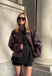 Jachetă din piele burgundy cu un design structurat, cu mâneci largi. Top negru cu guler înalt și pantaloni scurți completează ținuta. Fundal simplu.