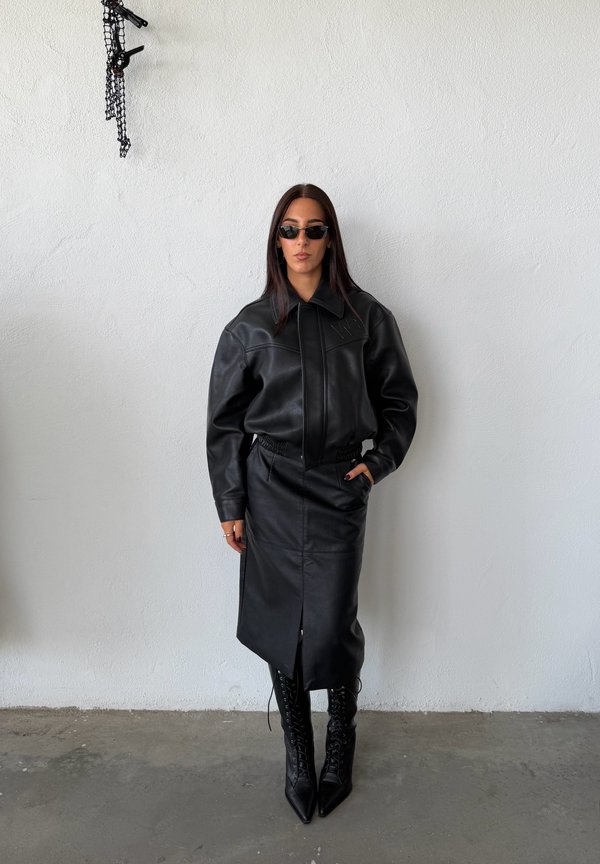 Femme portant une veste en cuir noire, une jupe midi en cuir noire, des bottes noires à lacets pointues et des lunettes de soleil foncées, debout contre un mur blanc.