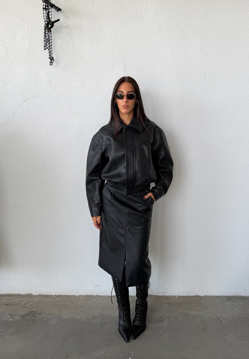Woman wearing black leather jacket, black leather midi skirt, black lace-up pointed boots, and dark sunglasses standing against white wall.