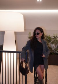 Kobieta w szarym, oversize'owym żakiecie, czarnej sukience i wysokich kozaczkach trzyma małą brązową torbę, a na jej twarzy znajdują się okulary przeciwsłoneczne oraz wyraźny pierścionek.