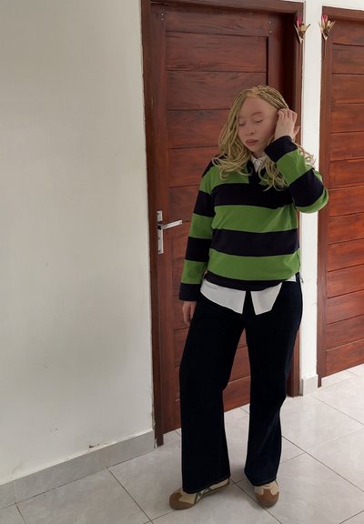 Camisa de manga larga a rayas verdes y negras sobre una camisa blanca con cuello, combinada con pantalones anchos negros y zapatos color marrón, de pie en interiores.