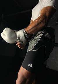 Baltas boksa cimdi ar teksturētu virsmu, apvienoti ar tumši melniem šortiem, kam ir zirnekļa tīkla dizains un logo akcenti, valkāti virs pieguļošiem melniem �šortiem.