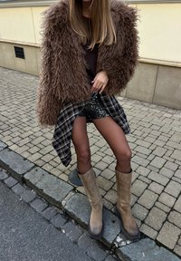 Personne portant une veste brune à poils longs, un haut noir, une chemise à carreaux nouée autour de la taille, un short à paillettes, des collants translucides et des bottes beige montantes jusqu'aux genoux sur le trottoir.