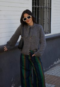 Cardigan gris de punto con detalle de lazo y botones, combinado con pantalones de pierna ancha a rayas multicolores. Bolso negro tipo bandolera y gafas de sol de gran tamaño.