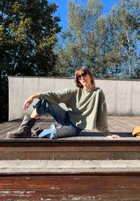 Top oversized verde-oliva de mangas compridas, jeans rasgados azul claro, botas de couro cinza com fivelas, em um deck de madeira, contra um céu azul.