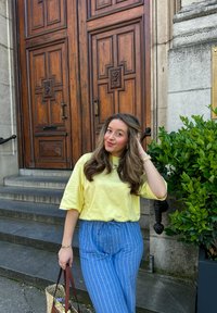 Jaune chemise à manches courtes, pantalon rayé bleu clair avec cordon de serrage, tenant un sac à main marron ; se tenant devant des portes en bois et des feuillages.
