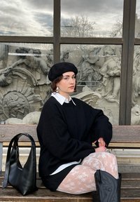 Svart oversized tröja, vit kragblus, genomskinliga blommiga strumpbyxor och svarta cowboyboots. En svart handväska vilar på en träbänk.