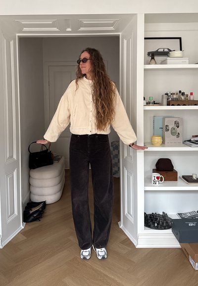 Mujer con cabello largo y rizado, que lleva gafas de sol, chaqueta crema y pantalones negros, de pie en una puerta con los brazos extendidos, estanterías con artículos a la derecha.