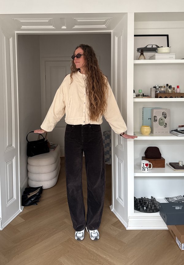 Donna con lunghi capelli ricci che indossa occhiali da sole, giacca color crema e pantaloni neri, in piedi nella porta con le braccia estese, scaffali con oggetti alla sua destra.