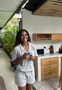 Femme souriante en pyjama gris tenant une tasse bleue, debout dans une cuisine avec des armoires en bois et des plantes d'intérieur visibles à l'extérieur.