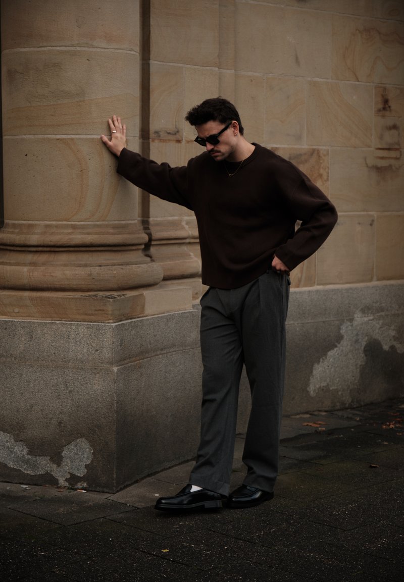 Tamnosmeđi vuneni džemper, sive plisirane hlače, crne kožne cipele s bijelim čarapama, stojeći pokraj kamenog stupa uz teksturirani zid.