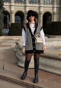 Czarnobiała kamizelka w wzór houndstooth z dużymi guzikami, noszona na białej koszuli. Dodatki obejmują małą torebkę w jasnym kolorze i czarne botki.
