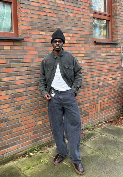 Chaqueta de tela oscura con dos bolsillos en el pecho, llevada sobre una camisa blanca, combinada con pantalones gris holgados y zapatos marrones, frente a una pared de ladrillo.