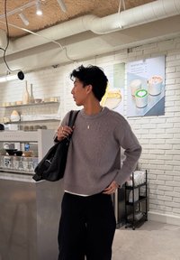 Άτομο με γκρι πουλόβερ που κρατά μαύρη τσάντα κοιτάζει προς το μενού του καφέ στον λευκό τοίχο από τούβλα πίσω από τον πάγκο με ποτήρια και εξοπλισμό για καφέ.