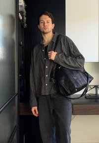 Homme portant une chemise à carreaux noir et blanc et un jean foncé, portant un grand sac à bandoulière en cuir noir à l'intérieur.