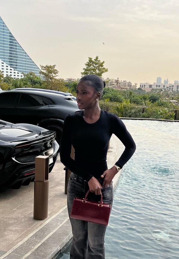 Femme en haut noir et jean tenant un sac à main rouge debout près d'une piscine avec des voitures de luxe et de la verdure en arrière-plan.