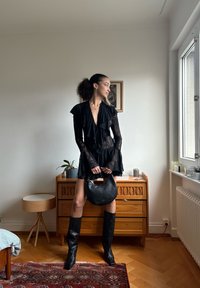 Robe en dentelle noire avec volants, manches longues et profond décolleté en V. Le mannequin tient un sac hobo noir brillant, assorti à des bottes noires montant jusqu'aux genoux.