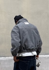 Siva, teksturirana bomber jakna s visokim ovratnikom i elastičnim rubom. Na stražnjoj strani ima oznaku i nosi se preko tamnih traperica.