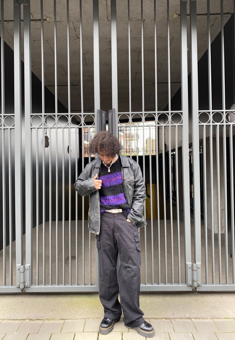 Fekete bőrkabát egy lila és fekete csíkos bolyhos pulcsi fölött, fekete cargo nadrággal és nagy talpú csizmával párosítva, egy kerítéssel ellátott háttér előtt.