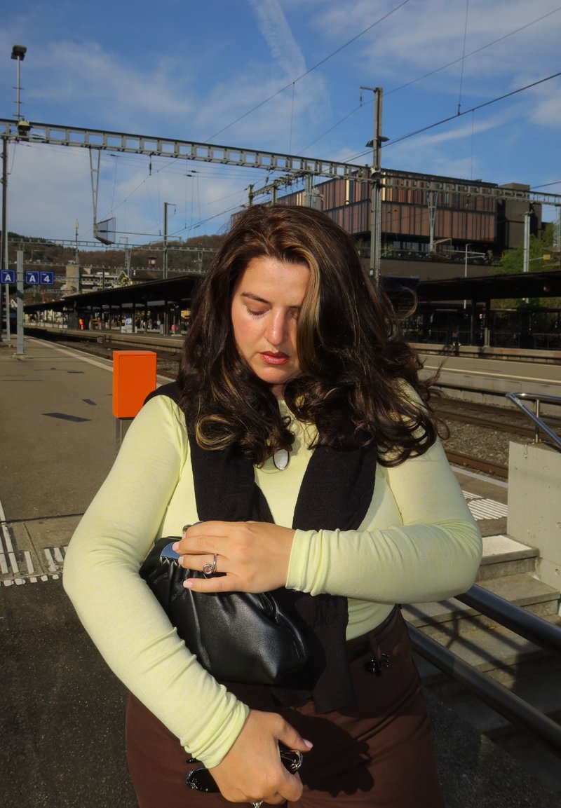 Femme aux cheveux foncés portant un haut à manches longues jaune et une écharpe noire, tenant un sac noir sur un quai de gare en plein air.