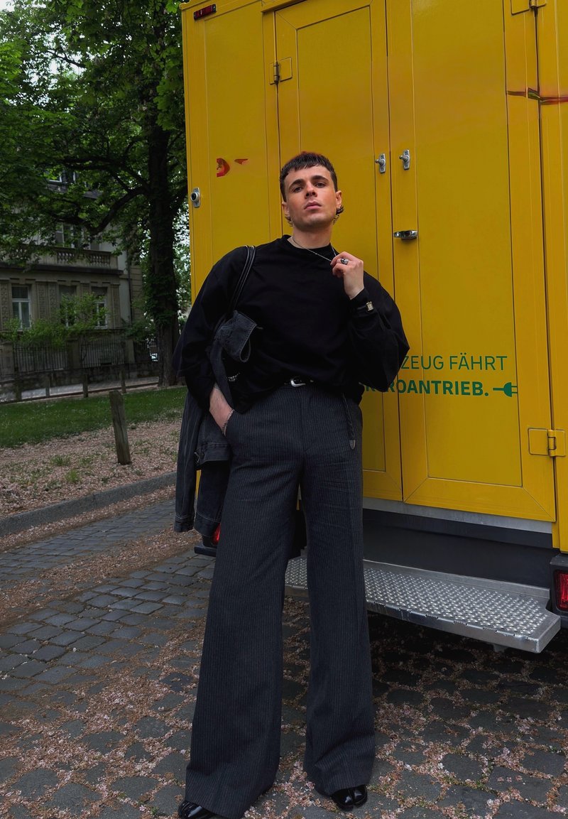 Junger Mann in schwarzem Top und weitem Hosen lehnt an einem gelben Fahrzeug mit grünem Text in einem blätterreichen urbanen Park.
