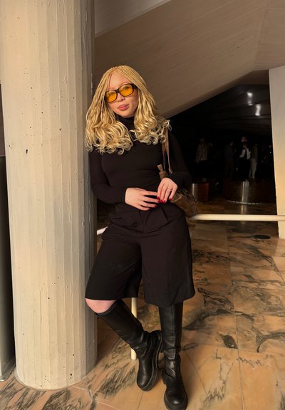Jersey de cuello alto negro y pantalones cortos, botas negras hasta la rodilla, gafas de sol doradas y cabello rubio ondulado. De pie cerca de un pilar gris en un espacio interior.